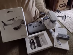 A white DJI Mini drone, its controller, and accessories are laid out on a textured surface, possibly a couch or carpet. The drone is in the center, with its propellers folded. To the left, a white box with an image of the drone is visible. A controller with joysticks and buttons is positioned behind the drone. Various accessories, including batteries, cables, and a gimbal protector, are neatly arranged in open boxes in front and to the right of the drone. The lighting is soft, highlighting the details of the products. The overall scene suggests a new product unboxing or preparation for flight.