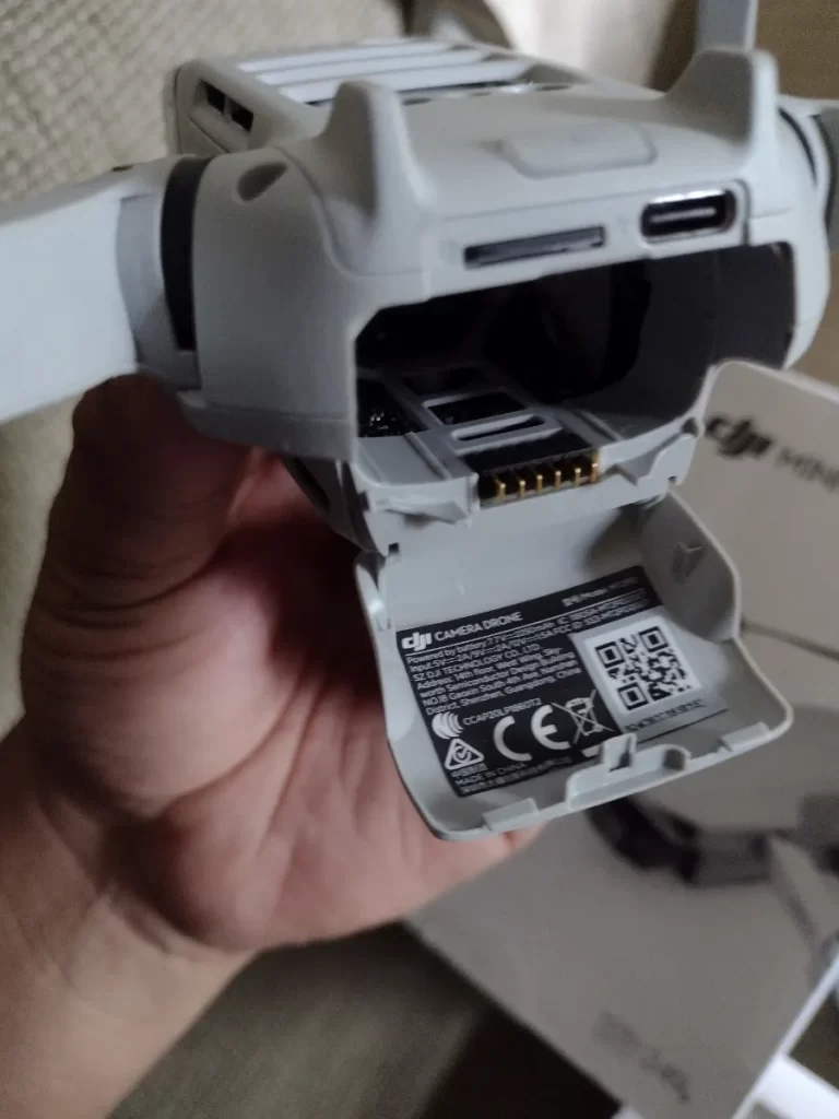 A close-up, slightly low-angle shot shows a person's hand holding a white drone. The drone's battery compartment is open, revealing the battery and some electronic components inside. The hand is visible in the foreground, with the thumb and index finger gently holding the drone. The drone is primarily white with some black accents. In the background, a blurred white object, possibly a box, is visible. The lighting is relatively soft, highlighting the details of the drone and the hand. The image has a shallow depth of field, focusing on the drone.