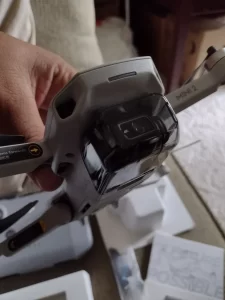 A close-up, slightly blurry shot shows a person's hand holding a gray and black drone. The drone's body is predominantly gray, with a glossy black camera module integrated into the front. The camera lens is dark and reflective. The drone's arms are extended, and the propellers are visible. The background is out of focus, with hints of white packaging and a wooden surface. The lighting appears to be indoor, with soft shadows. The image has a shallow depth of field, emphasizing the drone in the foreground.
