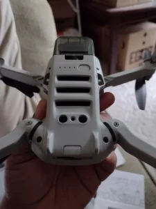 A close-up, slightly blurry shot shows a person's hand holding a gray and black drone. The drone's body is predominantly gray, with a glossy black camera module integrated into the front. The camera lens is dark and reflective. The drone's arms are extended, and the propellers are visible. The background is out of focus, with hints of white packaging and a wooden surface. The lighting appears to be indoor, with soft shadows. The image has a shallow depth of field, emphasizing the drone in the foreground.