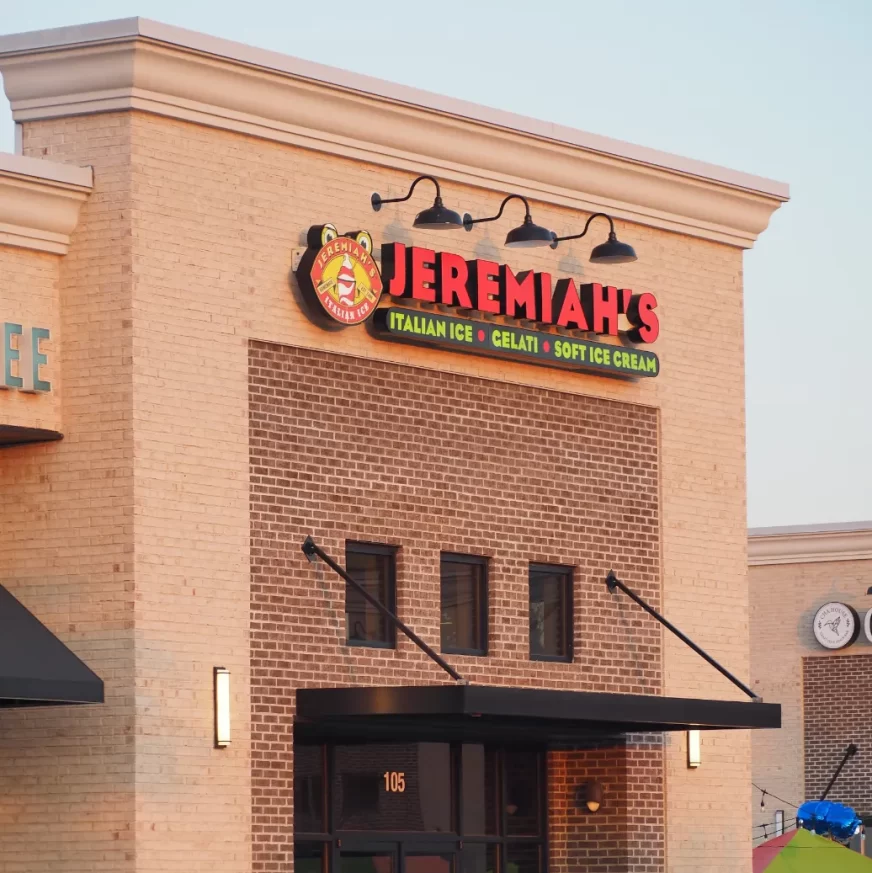 A close-up, eye-level shot captures the facade of a building. The main focus is a prominent sign with the name "JEREMIAH'S" in large, illuminated red letters. Above the name, a circular logo features a cartoonish pig character with a grin. Below "JEREMIAH'S," smaller text reads "ICE CREAM • GELATO • SORBET." The sign is mounted on a brick wall composed of light brown bricks arranged in a stretcher bond pattern. Four gooseneck light fixtures with exposed bulbs hang above the sign, casting a warm glow.