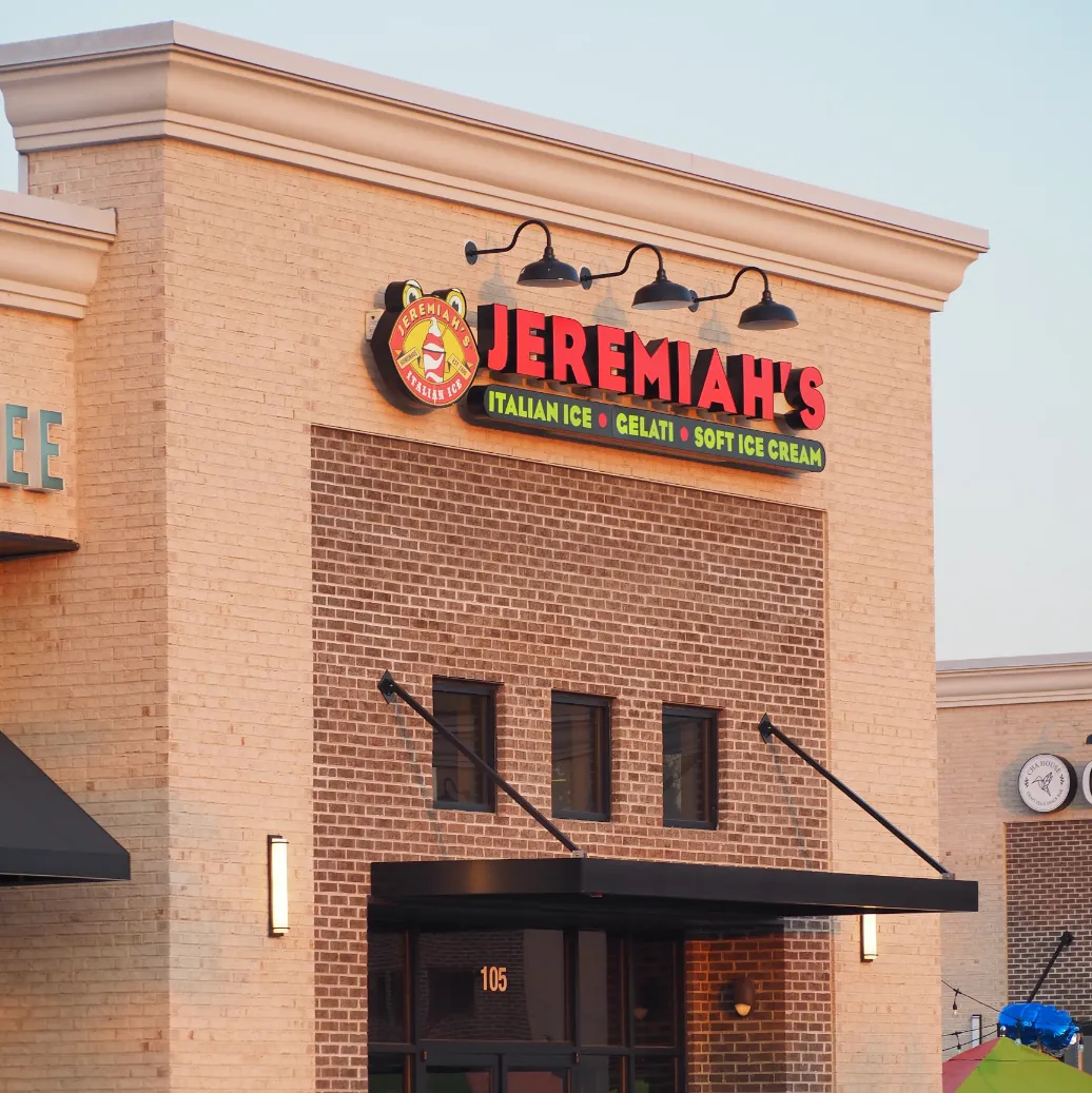 A close-up, eye-level shot captures the facade of a building. The main focus is a prominent sign with the name "JEREMIAH'S" in large, illuminated red letters. Above the name, a circular logo features a cartoonish pig character with a grin. Below "JEREMIAH'S," smaller text reads "ICE CREAM • GELATO • SORBET." The sign is mounted on a brick wall composed of light brown bricks arranged in a stretcher bond pattern. Four gooseneck light fixtures with exposed bulbs hang above the sign, casting a warm glow.