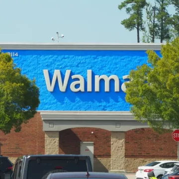 large white letters against a vibrant blue background, the walmart branding on a building, in the foreground are trees and cars, the name is slightly obscured by foliage