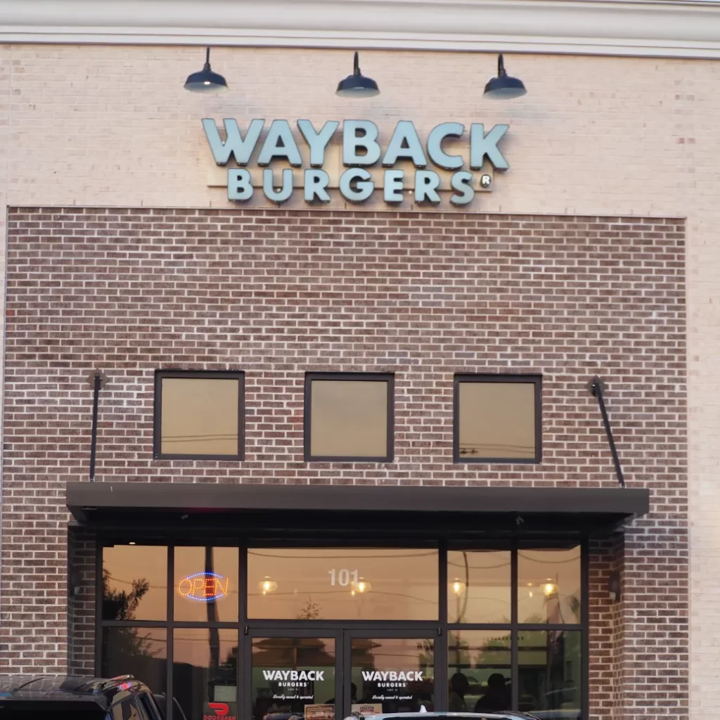 A close-up, eye-level shot of the exterior of a Wayback Burgers restaurant. The building is constructed of rough, reddish-brown brick with visible mortar lines. Above the entrance, a dark awning covers large, reflective glass windows. Three identical square windows with golden-tinted glass are positioned above the awning, spaced evenly apart. Directly above these windows, a prominent sign spells out "WAYBACK BURGERS" in illuminated, teal-colored block letters. Three black, gooseneck-style outdoor lights are mounted on the building's facade above the sign, casting a subtle downward light. The overall impression is one of a clean, modern fast-casual dining establishment.