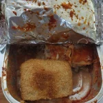Takeout foil container with lasagna covered in marinara sauce and a sesame seed bread roll