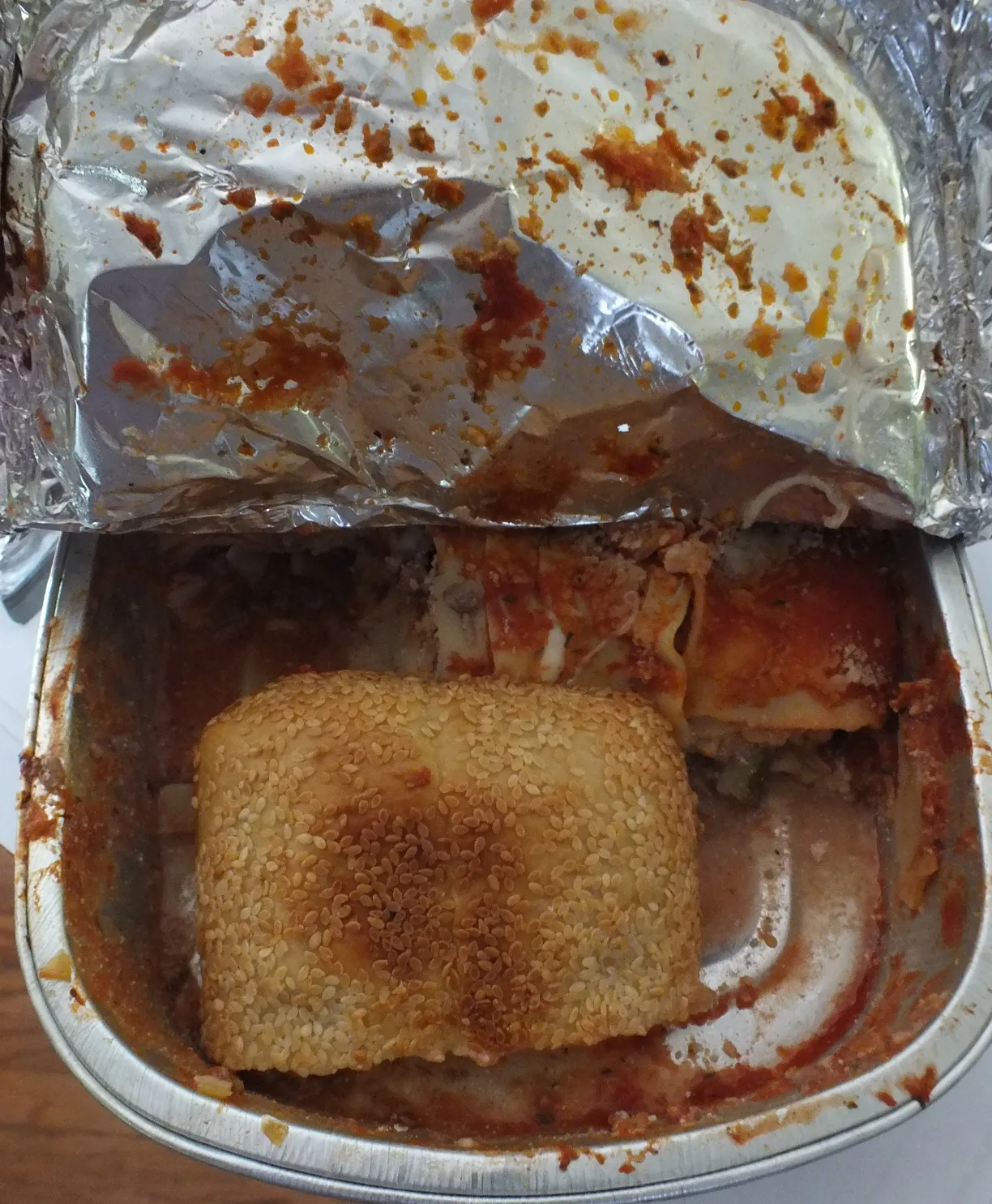 Takeout foil container with lasagna covered in marinara sauce and a sesame seed bread roll