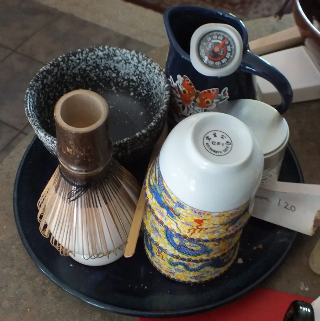 Matcha tea preparation set on a tray with a bamboo whisk, scoop, speckled bowl, stacked cups, and pitcher with thermometer