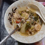 Fork lifting flat noodles from a bowl of soup with leafy greens, mushrooms, and a slice of red chili