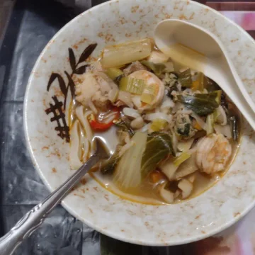 Fork lifting flat noodles from a bowl of soup with leafy greens, mushrooms, and a slice of red chili