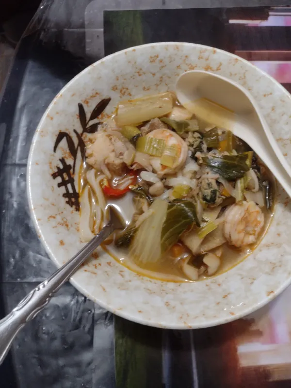 Fork lifting flat noodles from a bowl of soup with leafy greens, mushrooms, and a slice of red chili