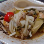 Fork lifting flat noodles from a bowl of soup with leafy greens, mushrooms, and a slice of red chili