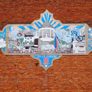 Colorful mural on a red brick wall reading Wake Forest, featuring local landmarks, a steam train, and community symbols