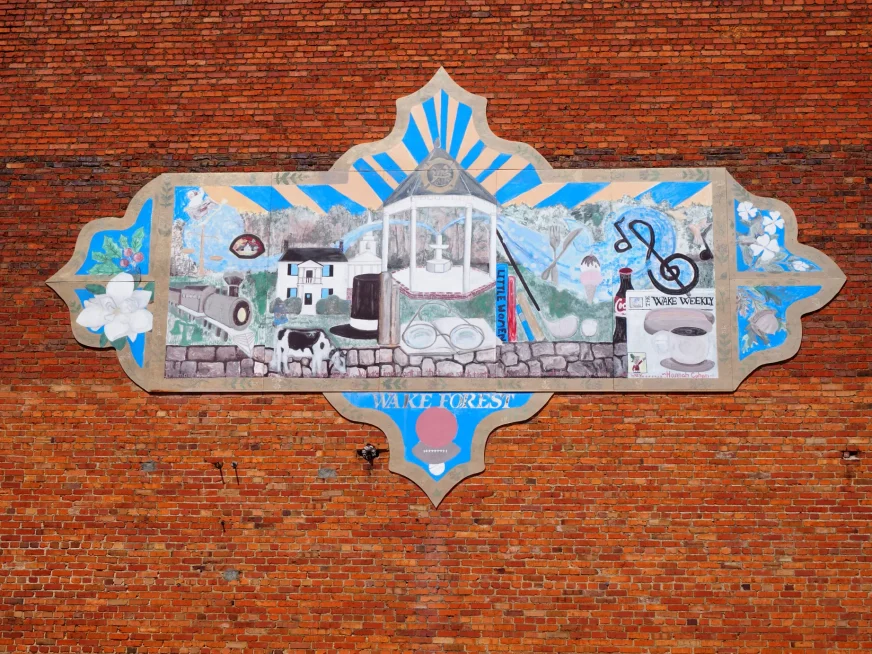 Travel Through Sprawling Wake Forest, NC Colorful mural on a red brick wall reading Wake Forest, featuring local landmarks, a steam train, and community symbols